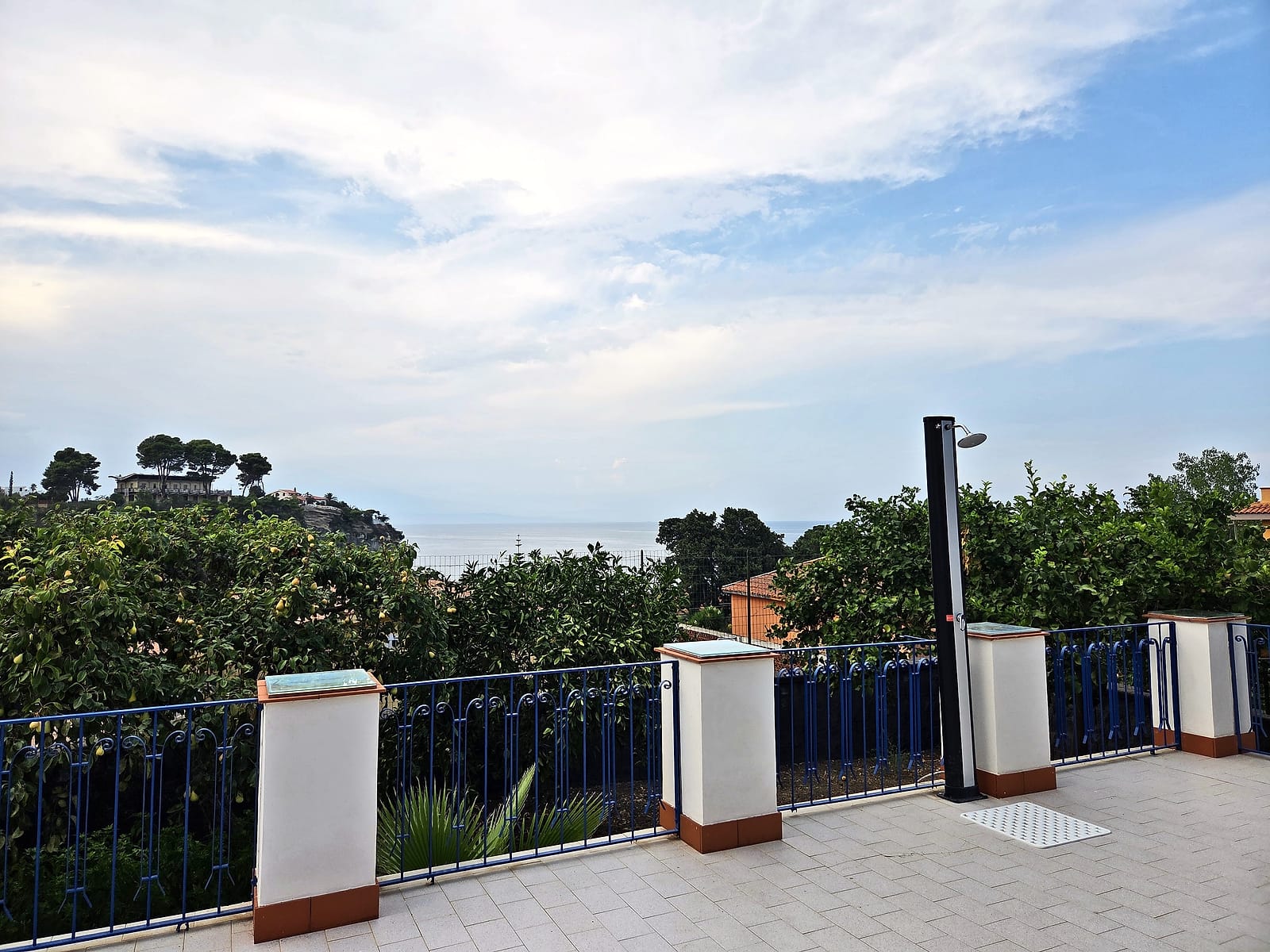 La terrazza con la doccia esterna, la ringhiera blu e uno scorcio di mare oltre gli alberi